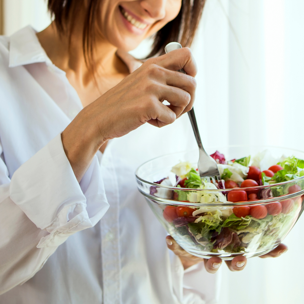 A woman smiling and eating from a bowl of greens and vegetables, demonstrating the importance of nutrient-dense foods in a detox for moms in menopause.