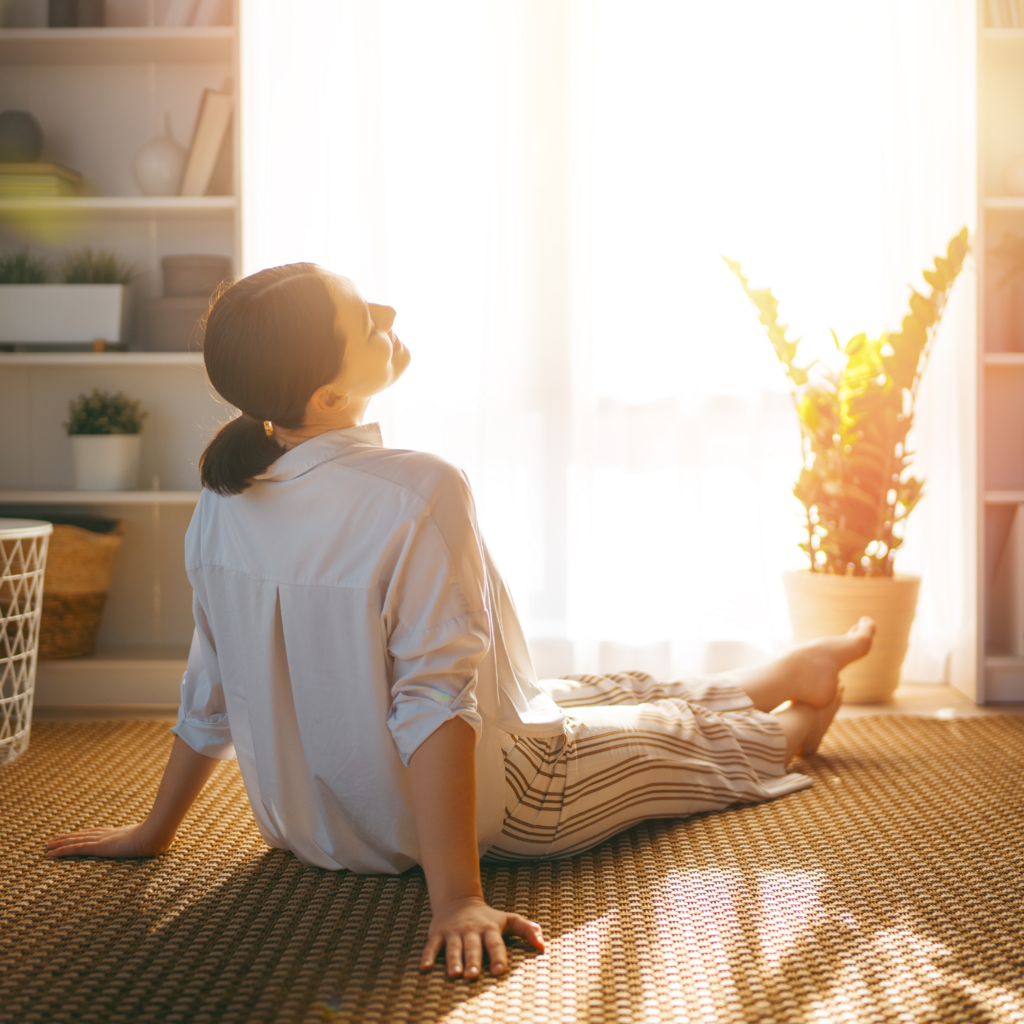 A woman relaxing happily on the floor facing the window in a sun-filled room, illustrating the importance of prioritizing rest in detox for moms in menopause.