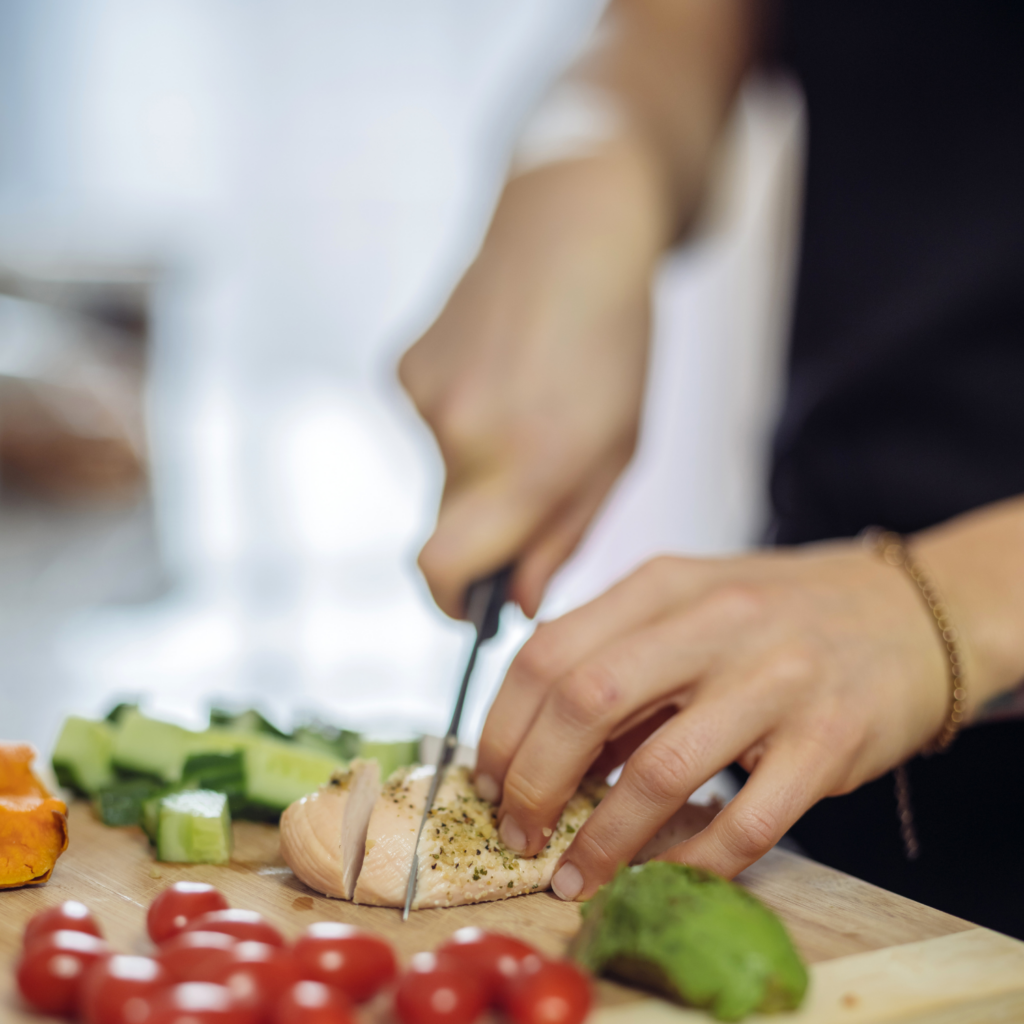 A woman preparing food in her kitchen, slicing chicken breast with vegetables on the counter, addressing how to simplify meal prep during a detox for moms in menopause.