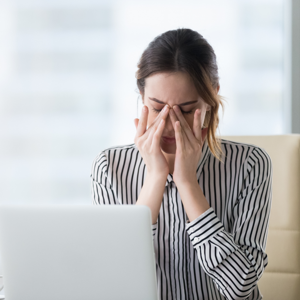 A woman sitting in front of a computer, massaging her sinus area in discomfort, highlighting signs that detox may help moms in menopause.