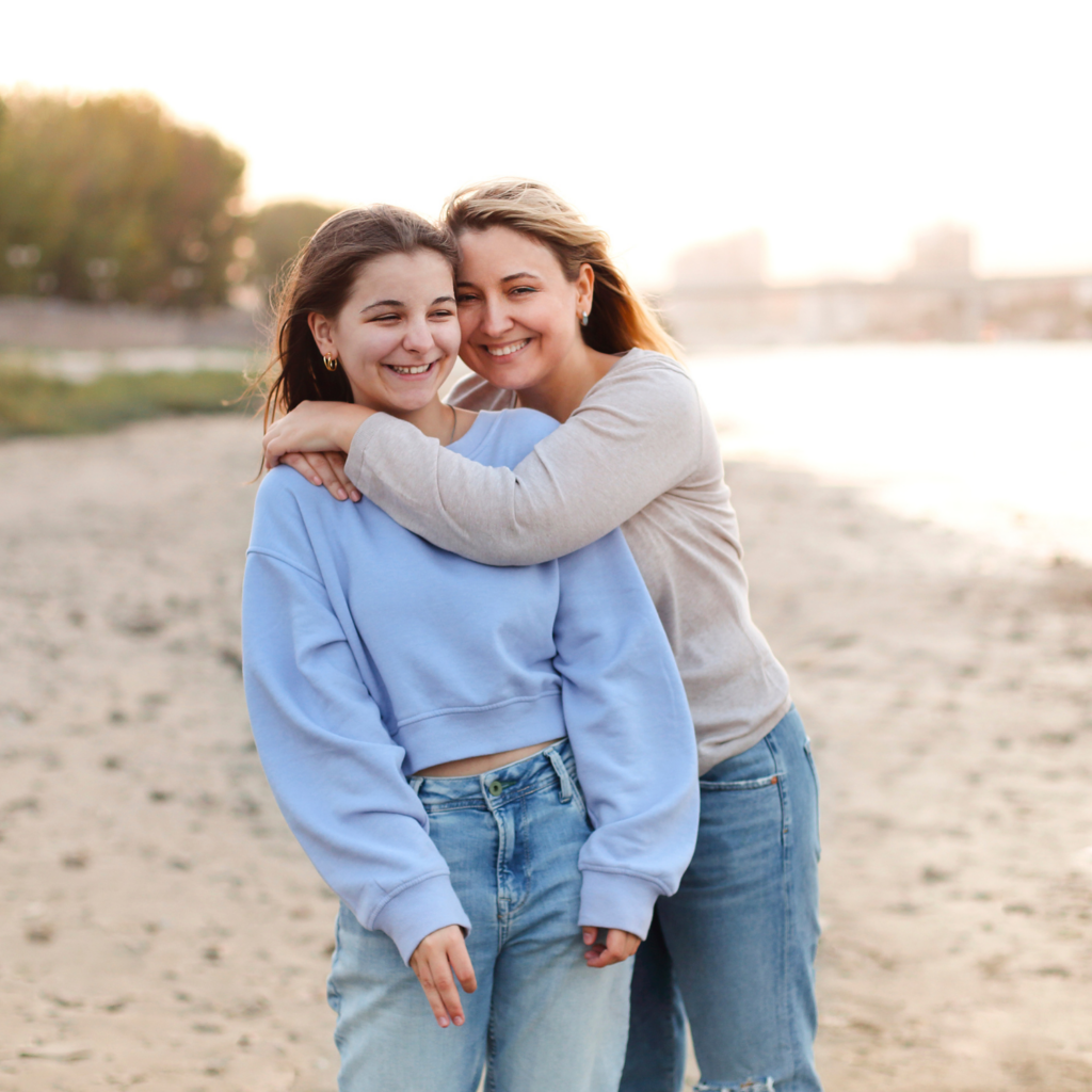 A mom smiling and hugging her teenage daughter on the beach, symbolizing the positive effects of detox for moms in menopause.