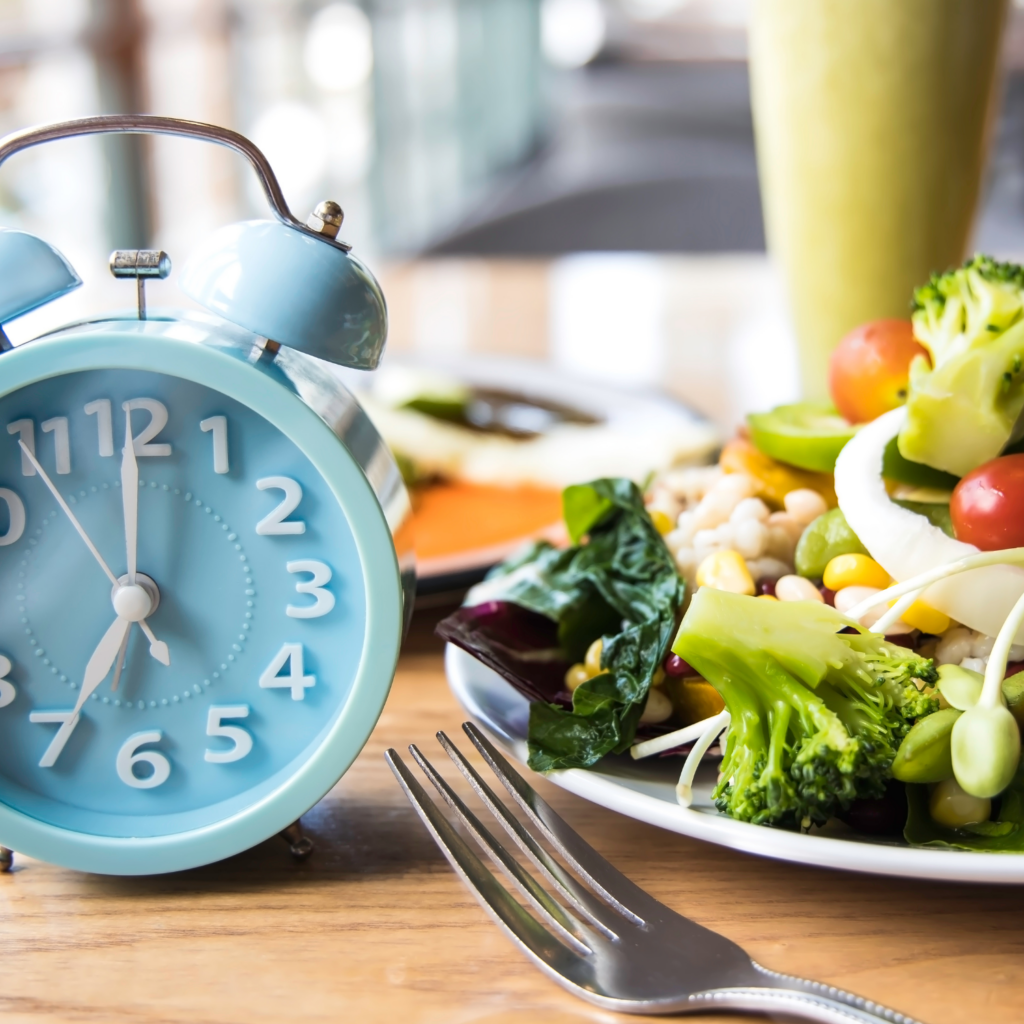 A blue clock on a breakfast table with a plate of healthy food, representing strategic fasting for energy and hormones.